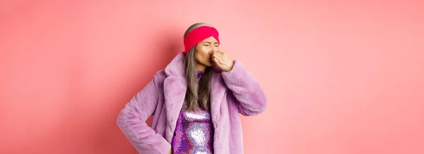 Portrait of stylish old asian woman in fashionable purple coat and dress, shut nose from bad smell, express disgust and aversion as something sink awfully, standing over pink background.