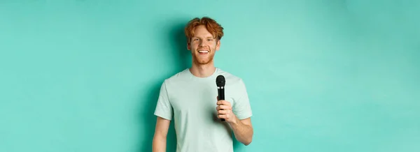 Young redhead man with beard, wearing t-shirt, holding microphone and making speech, singing karaoke, standing over mint background.