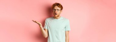 Gözlüklü, kafası karışık, ağzı açık, kameraya bakan, hiçbir şey anlamayan, pembe arka planda duran yakışıklı kızıl adamın portresi..