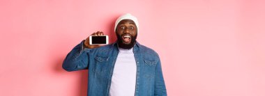 Çevrimiçi alışveriş ve teknoloji kavramı. Şaşırmış siyah adam telefon ekranını yatay olarak gösteriyor ve kameraya bakıyor heyecanlı, pembe arka planda duruyor..