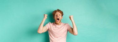 Yakışıklı kızıl adam maç seyrediyor ve tezahürat yapıyor, bağırıyor ve yumruk pompalıyor, ekrana bakıyor ve yarışmayı seyrediyor, takımı destekliyor, turkuaz arka planda duruyor..