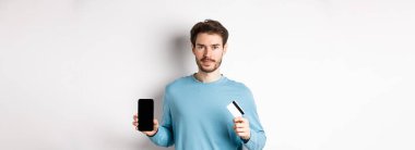 Beyaz arka planda duran, boş akıllı telefon ekranı ve plastik kredi kartı gösteren yakışıklı beyaz bir adam..