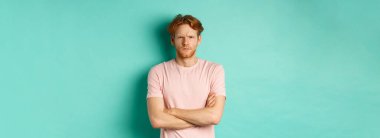 Kızıl saçlı ve sakallı genç adam gücenmiş görünüyor, aşağılanmış ve kameraya bakıyor, sana somurtuyor, kollarını göğsüne dayayıp savunma yapıyor, turkuaz arka planda duruyor..