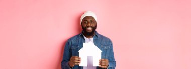 Emlak konsepti. Mutlu gülümseyen Afro-Amerikalı adam ev modelini tutuyor, daire alıyor, pembe arka planda duruyor..