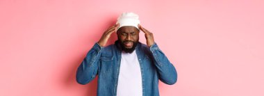 Sıkıntılı siyah adam baş ağrısı çekiyor, başıyla temas ediyor ve endişeli bir yüz ifadesiyle yüzünü buruşturuyor, pembe arka planda duruyor..