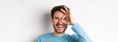 Heyecanlı gülümseyen adam, tamam tabelasından bakıyor. Mutlu yüz, onay ve tanıtım teklifi gibi beyaz arka planda duruyor..