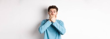 Şok olmuş genç bir adamın elleri ile ağzını kapayarak bakması, dedikoduları duyması, beyaz arka planda durması..