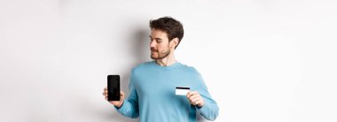 Mobil bankacılık. Plastik kredi kartı gösteren yakışıklı beyaz adam ve beyaz arka planda duran boş mobil ekrana bakıyor..