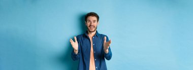 Neşeli genç adam kameraya el kaldırıyor ve gülümsüyor, senin için seviniyor, iyi iş çıkarıyor, mavi arka planda duruyor..