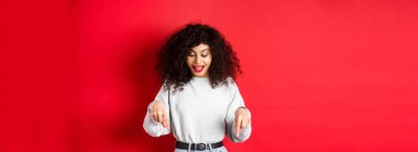 Kıvırcık saçlı rüya gibi bir kadın, işaret ediyor ve aşağı bakıyor, heyecanlı, tanıtıma bakıyor, kırmızı arka planda duruyor..