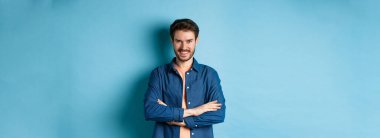 Kendine güveni tam genç bir adam kendini profesyonel hissediyor, kollarını göğsünde kavuşturuyor ve sırıtıyor, mavi arka planda duruyor..