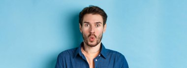 Şaşırmış ve şaşırmış bir adamın yakın portresi. Vay canına, kameraya bakıyor, harika bir teklif sunuyor, mavi arka planda duruyor..