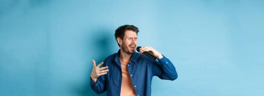 Romantik adam mikrofon ve jest ile şarkı söylüyor, karaoke yapan şarkıcı, mavi arka planda duruyor..