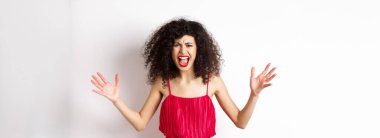 Kıvırcık saçlı kızgın genç bir kadın, kırmızı elbise giyiyor, çığlık atıyor ve tartışıyor, nefret ve öfkeyle bakıyor, beyaz arka planda duruyor..