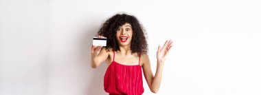 Alışverişe. Kırmızı elbiseli, şaşırtıcı kıvırcık saçlı kadın, plastik kredi kartı gösteriyor ve heyecandan çığlık atıyor, beyaz arka planda duruyor..