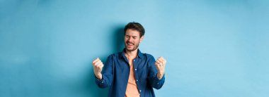 Başarıyı kutlayan mutlu bir adam tasviri, evet diye bağırıyor ve yumruklarını sallıyor, hedefe ulaşıyor ve başarıyor, kazanıyor ve dans ediyor, mavi arka planda duruyor..