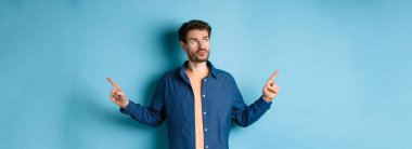 Kararsız bir adam günlük kıyafetlerini giyiyor, seçimler yapıyor, parmaklarını yana çeviriyor ve dalgın bakıyor, mavi arka planda duruyor..