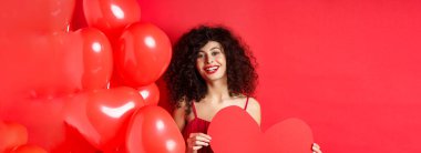 Sevgililer günün kutlu olsun. Gerçek aşkı bekleyen, büyük kırmızı kalbi olan ve kırmızı arka planda balonların yanında duran romantik bir kız..