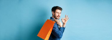 Omuzunda alışveriş çantası tutan yakışıklı genç adam, kameraya bakmak için arkanı dön ve iyi niyet göster, tavsiye dükkânı, mavi arka plan.