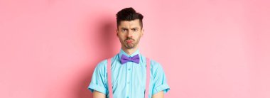 Üzgün ve gücenmiş genç bir adam, kaşlarını çatmış ve dudaklarını büzmüş, birine kızgın bakıyor, papyonlu pembe arka planda hayal kırıklığına uğramış duruyor..