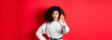 Kendine güveni tam bir kadın dur demek için elini uzatıyor, eylemi onaylamıyor ve yasaklıyor, hiçbir hareket yapmıyor, kırmızı arka planda duruyor ve bir şeyi yasaklıyor..