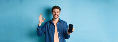 Yakışıklı genç adam iyi bir hareket ve boş akıllı telefon ekranı gösteriyor. Uygulama ya da şirket öneriyor. Mavi arka planda duruyor..