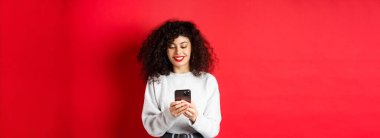 Gülümseyen Avrupalı kadın cep telefonu ekranı okuyor, akıllı telefondan mesaj yolluyor, kırmızı arka planda eşofman üstü duruyor..