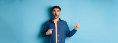 Şaşırmış ve şaşırmış beyaz adam hayranlıkla soluk soluğa kalmış, sol logoyu işaret ediyor, müthiş bir tanıtım yapıyor, mavi arka planda duruyor..