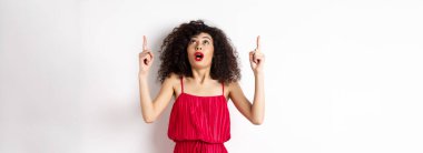 Gece makyajlı, kıvırcık saçlı, heyecanlı ve dik duran, reklam yapan, beyaz arka planda kırmızı elbiseli harika bir kız..