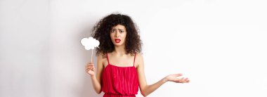 Kırmızı elbiseli kafası karışmış genç bir kadın, üzgün ve üzgün görünüyor, yorum bulutu çubuğu tutuyor, beyaz arka planda duruyor..