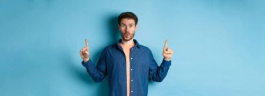 Şaşırmış genç adam diyor ki: Vay canına, reklamı kontrol ediyor, parmaklarını havaya kaldırıyor ve etkilenmiş görünüyor, mavi arka planda duruyor..