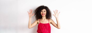 Kıvırcık saçlı, kırmızı elbiseli, on numarayı parmaklarıyla gösteren, beyaz arka planda duran güzel bir manken..