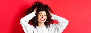 Yumuşak kıvırcık saçlara dokunan, gülümseyen, kırmızı arka planda duran kaygısız kadının yakın çekimi. Saç bakımı ve güzellik konsepti.