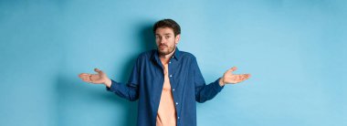 Kafası karışmış sakallı, omuzlarını silkmiş, kameraya habersiz bakan, hiçbir şey bilmeyen, mavi arka planda duran genç adam..