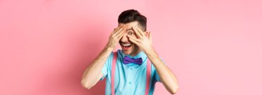 İlgi çekici papyonlu komik adam, parmaklarının arasından ilginç bir şeye bakıyor, havalı bir şeye bakıyor, pembe arka planda duruyor..