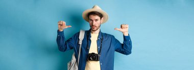 Yazlık şapkalı yakışıklı adam, böbürlenerek bakıyor, tatillerde seyahat ediyor, sırt çantası ve kamera taşıyor, mavi arka plan..