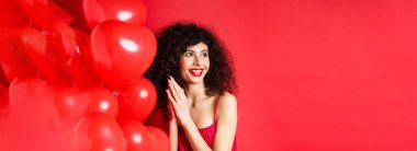 Heyecanlı kıvırcık saçlı güzel bir kadın, kalp balonlarının yanında duruyor ve avuç içlerini birbirine sürtüyor, iyi bir anlaşma ya da zevk bekliyor, kırmızı arka plan.