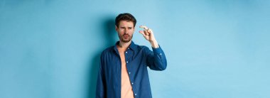 Kafası karışmış bir adam, küçük boyda, şüpheli ya da şüpheci görünüyor, mavi arka planda duruyor. Boşluğu kopyala