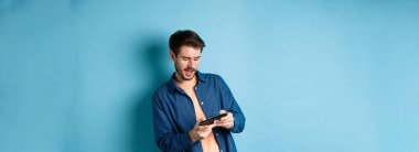 Genç adam cep telefonuyla video oyunu oynuyor, vücudunu eğiyor ve akıllı telefonu yatay olarak tutuyor, mavi arka planda duruyor..