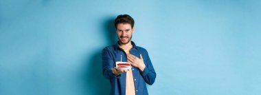 Kutlama ve tatil konsepti. Utanan ve doğum gününü kutlayan adam, yanan mumla doğum günü pastasını uzat, mavi arka planda dur..