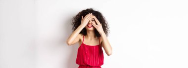 Kıvırcık saçlı, kırmızı elbiseli, elleriyle gözlerini kapatan ama parmaklarının arasından bakıp gülümseyen, ilginç, beyaz bir arka plana bakan komik bir genç kadın..