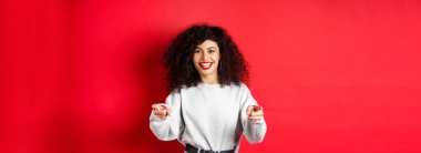 Kıvırcık saçlı neşeli kadın seni davet ediyor, yeni yetmeleri işe alıyor, kamerayı işaret ediyor ve gülümsüyor, iyi işi övüyor, kırmızı arka planda duruyor..