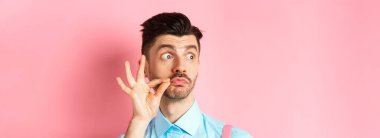 Genç beyaz adamın bıyıklara ve büzüşmüş dudaklara dokunup soldan logoya bakıp pembe arka planda durduğu yakın plan..