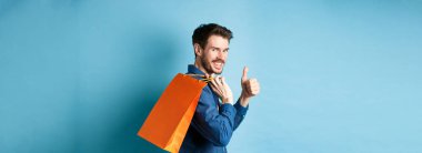 Omzunda turuncu alışveriş çantası tutan neşeli alışverişçi, kameraya doğru dön başparmaklar yukarı, dükkan tavsiye, mavi arka plan.