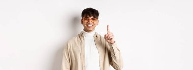 Gözlüklü yakışıklı beyaz adam bir parmağını gösteriyor ve gülümsüyor, beyaz arka planda duruyor..