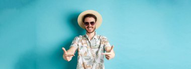 Yaz şapkalı ve Hawaii gömlekli mutlu bir turist. Ortadaki logoyu işaret ediyor. Bir şey gösteriyor, mavi arka planda duruyor..