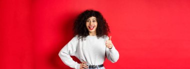 Kıvırcık saçlı mutlu bir genç kadın iyi iş çıkarmış, aferin deyin ve başparmağınızı kaldırın, onaylayın ve övün, kırmızı arka planda durun..