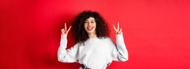 Kıvırcık saçlı şık bir genç kadın, gülümseyen mutlu ve barış işaretleri gösteren kırmızı arka planda sıradan bir kazak giyen..
