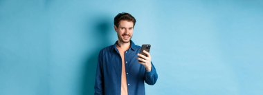 Çekici beyaz adam cep telefonu kullanıyor, mutlu görünüyor ve kameraya gülümsüyor, mavi arka planda duruyor..