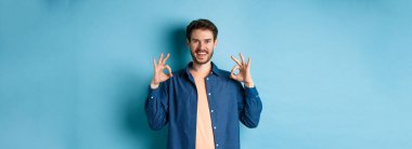 Tatmin olmuş genç adam gülümsüyor ve iyi bir jest yapıyor, iyi bir şeyi onaylıyor, mükemmel bir şeyi övüyor, mavi arka planda duruyor..
